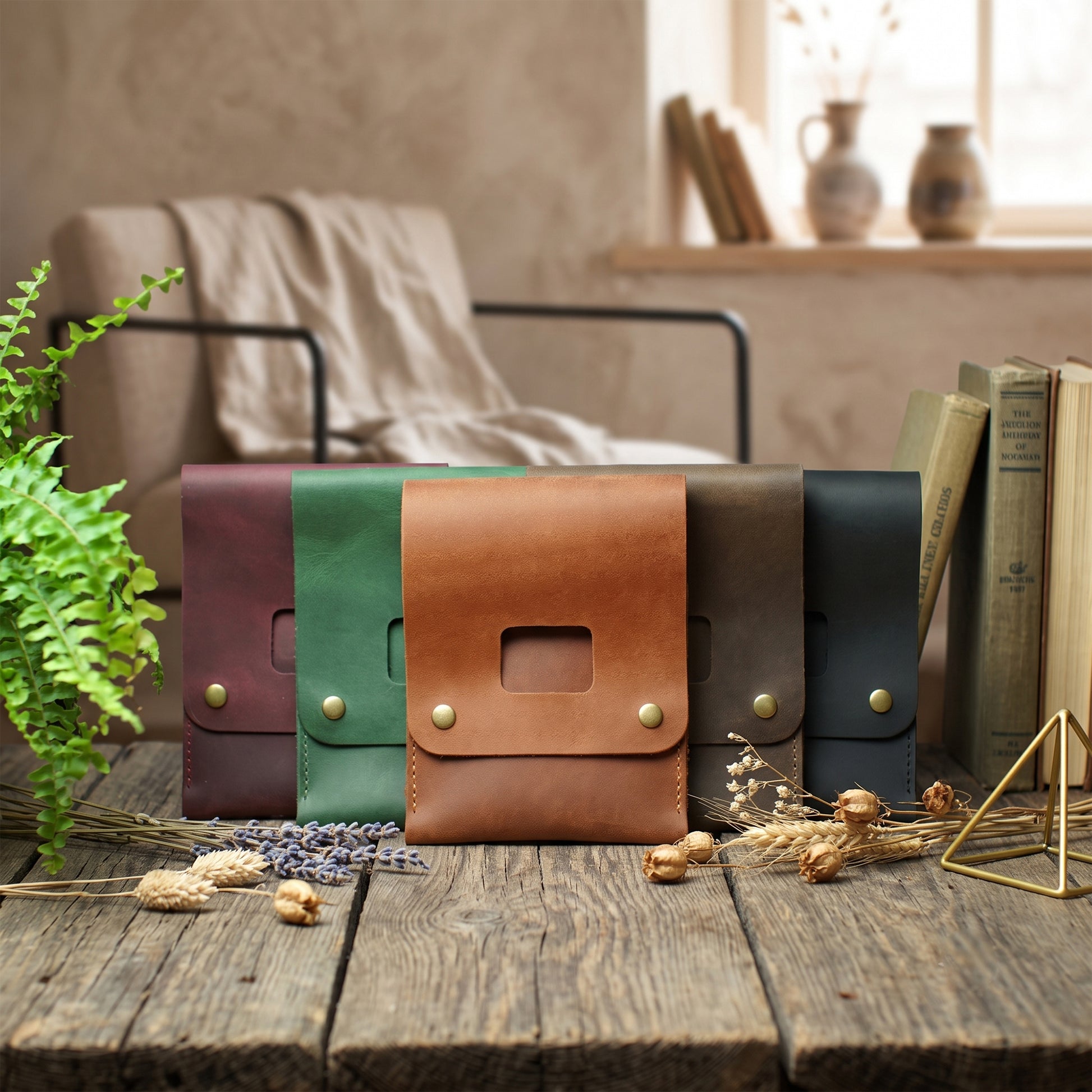 Five leather clutch or wallet-style accessories in different colors are arranged on a rustic wooden table in a styled interior setting with books, a chair, and dried flowers.