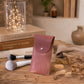A dusty rose leather makeup pouch with a silver snap button stands on a rustic wooden table surrounded by makeup brushes, dried flowers, and decorative books with warm fairy lights in the background.