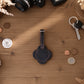 A dark brown leather luggage tag or key fob with a scalloped edge and a short strap, laid flat on a wooden tabletop surrounded by small props including keys, coins, a camera, and dried flowers.