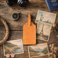 A tan leather luggage tag with a blank center panel, styled on a rustic wooden tabletop with vintage postcards, a compass, a camera, coins, rope, and a passport.