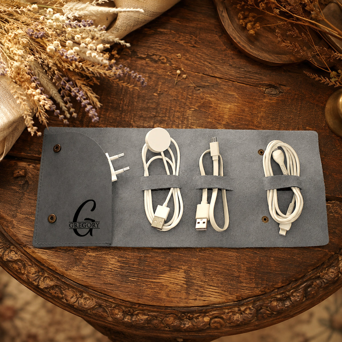 A gray roll-up pouch organizer laid open on a rustic wooden table, holding several white charging cables and a round white accessory. Dried flowers and a wooden bowl are visible around it.