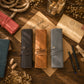 Three personalized leather wallet roll-up cases in dark brown, tan, and slate blue, displayed on a rustic wooden background with vintage-style stationery and decorative props.
