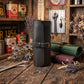 A black leather rolled canvas roll with a buckle strap stands on a rustic wooden table, surrounded by vintage-style tools, cans, dried flowers, and additional rolled leather goods in green and burgundy.