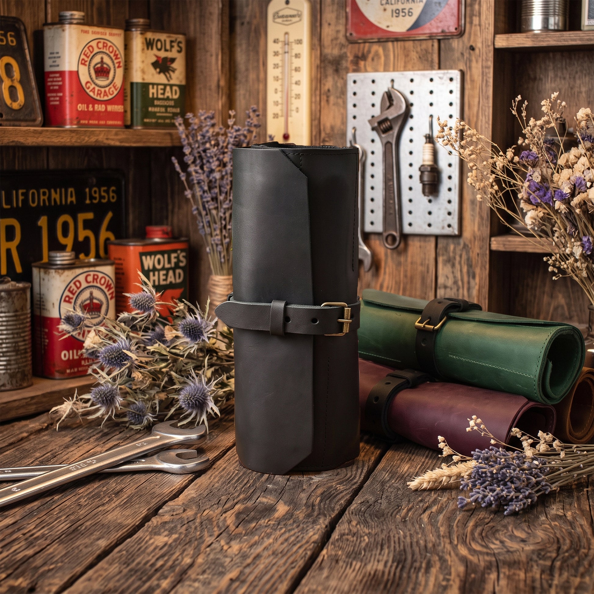 A black leather rolled canvas roll with a buckle strap stands on a rustic wooden table, surrounded by vintage-style tools, cans, dried flowers, and additional rolled leather goods in green and burgundy.