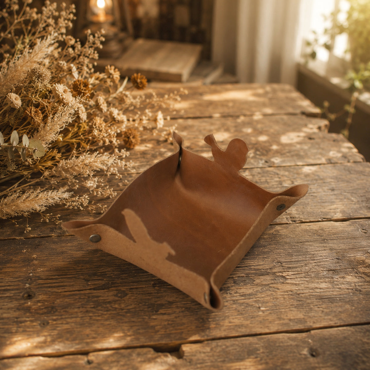 A brown leather tray with a deer head handle, resting on a rustic wooden table with dried flowers in the background.