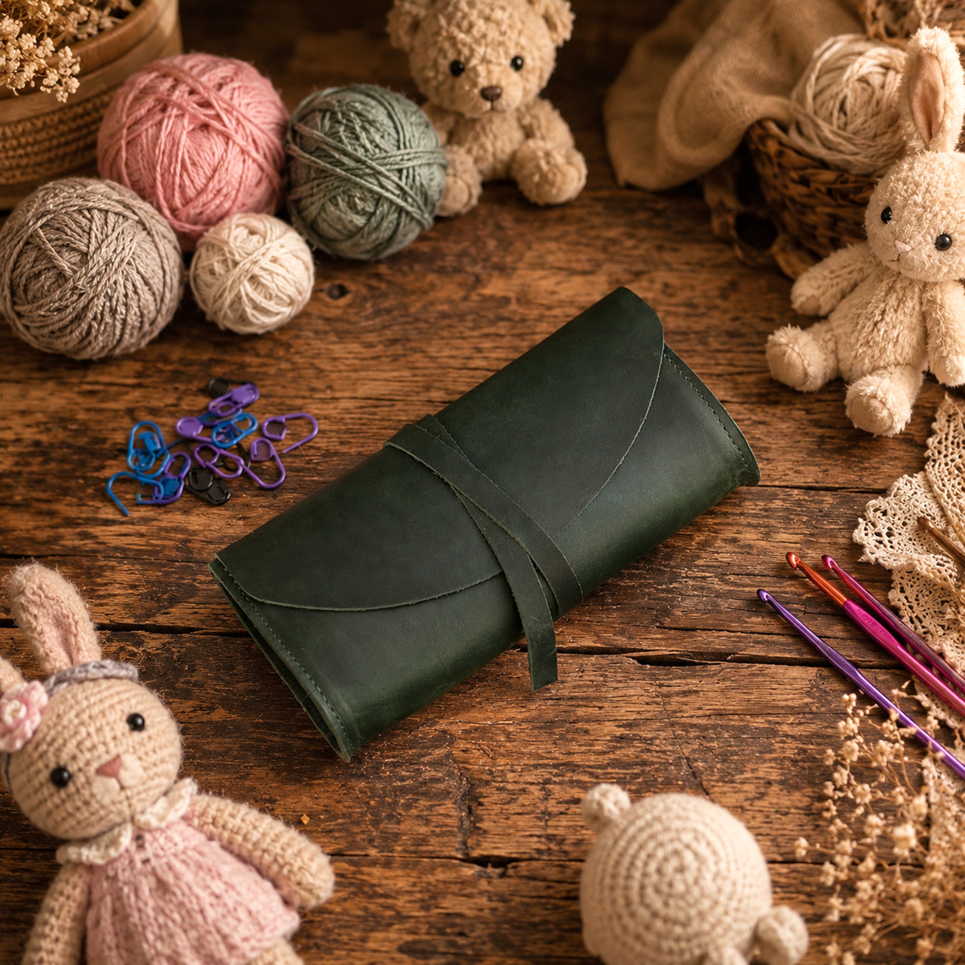 A dark green leather craft organizer or pouch with a wrap-around strap, styled on a rustic wooden table surrounded by crochet and knitting supplies, yarn balls, teddy bears, and knitted bunny dolls.