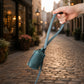 A hand holds up a small teal blue leather shoulder bag with a matching strap, set against a blurred cobblestone street at sunset.
