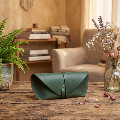 A green leather eyeglasses case with a flap closure and a small gold snap button, resting on a rustic wooden table in a cozy, warm-toned living room setting.