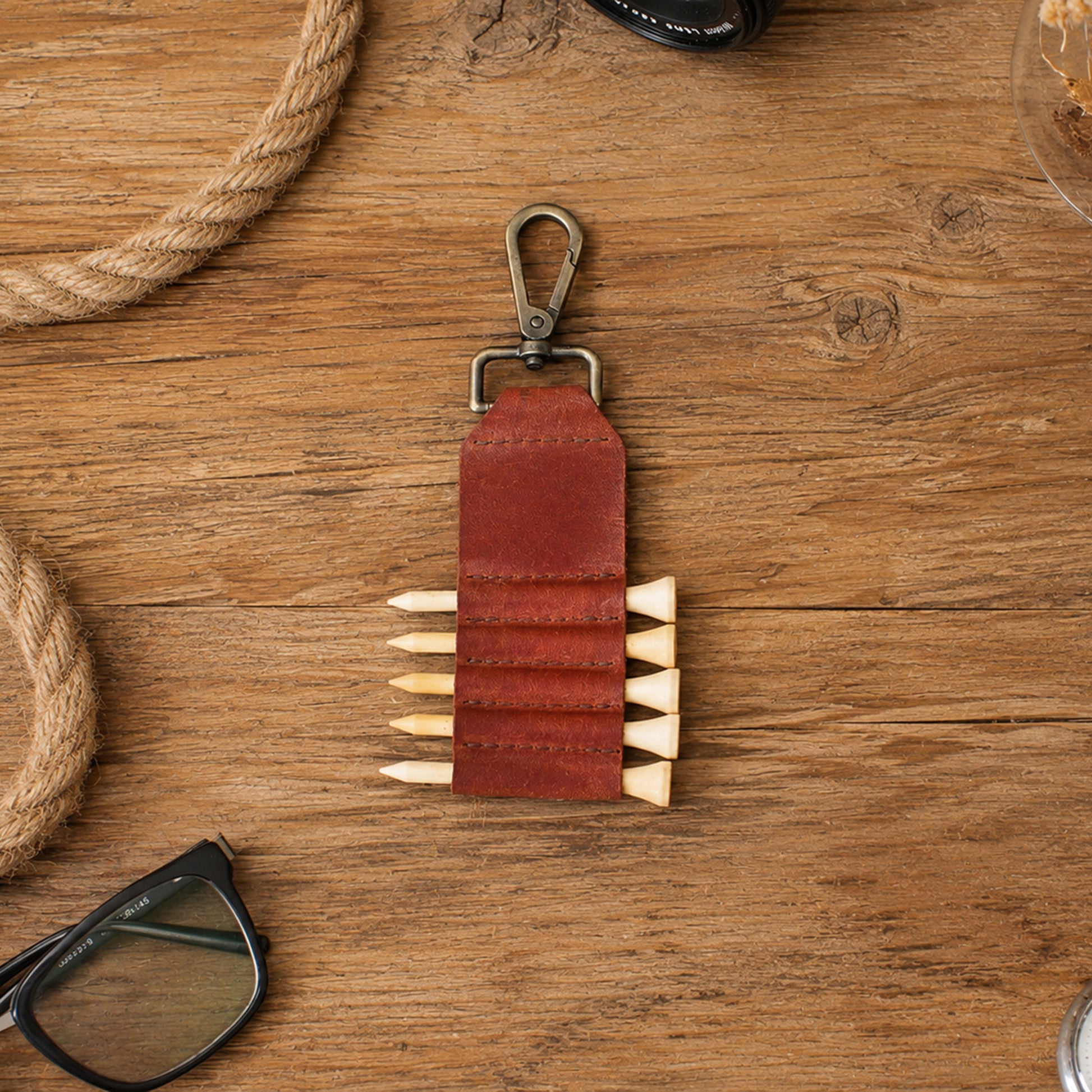 A brown leather keychain organizer with a metal clip and five elastic loops holding wooden pencils, displayed on a rustic wooden tabletop with rope and eyeglasses as props.
