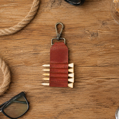 A brown leather keychain organizer with a metal clip and five elastic loops holding wooden pencils, displayed on a rustic wooden tabletop with rope and eyeglasses as props.