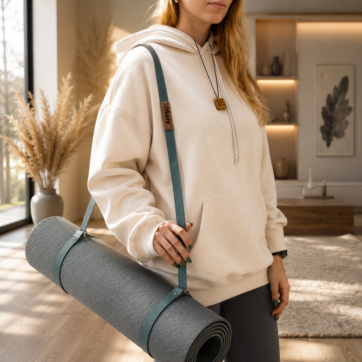 A woman in a cream hoodie carries a gray rolled yoga mat on a teal shoulder strap with a small wooden brand tag, standing in a bright, modern room.