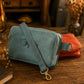 A dusty blue faux-leather crossbody bag with a black cursive script logo on the front, resting on a rustic wooden table next to an orange pouch.