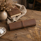A rolled brown leather journal or notebook cover tied with a matching strap, placed on a rustic wooden table. A closed brown notebook, a beige vase with dried flowers, a linen cloth, and a small dish with white earbuds are arranged around it in a styled setting.