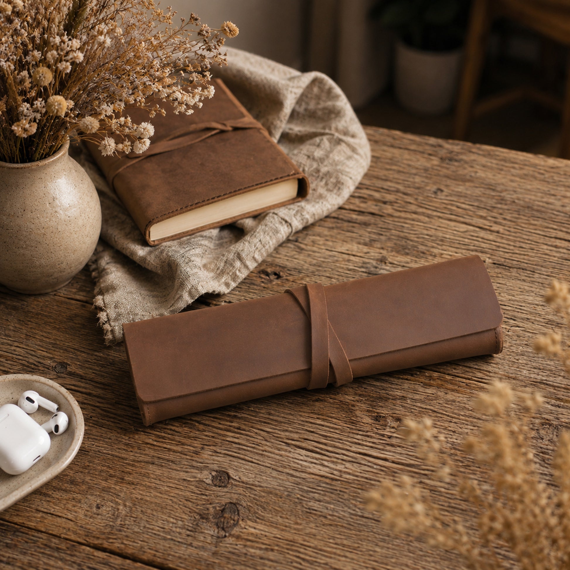 A rolled brown leather journal or notebook cover tied with a matching strap, placed on a rustic wooden table. A closed brown notebook, a beige vase with dried flowers, a linen cloth, and a small dish with white earbuds are arranged around it in a styled setting.
