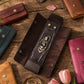 A dark brown leather jewelry organizer roll displayed open on a rustic wooden table, holding a silver chain necklace with a green and white stone pendant. Other leather jewelry pouches in brown, teal, tan, and magenta are arranged around it with pink tulips on the surface.
