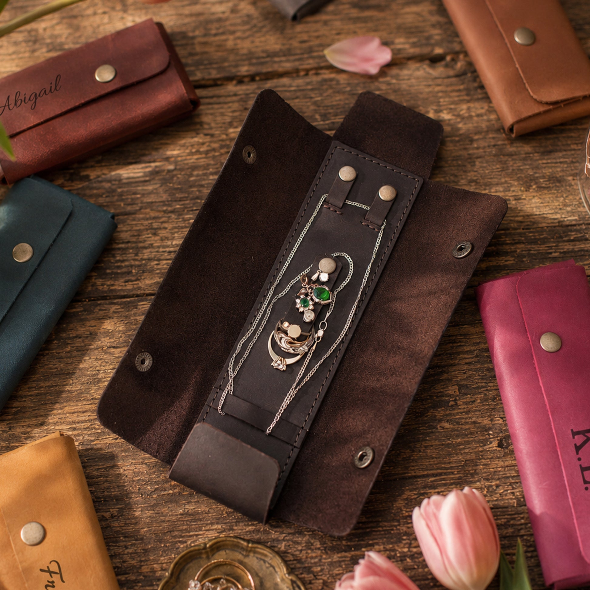 A dark brown leather jewelry organizer roll displayed open on a rustic wooden table, holding a silver chain necklace with a green and white stone pendant. Other leather jewelry pouches in brown, teal, tan, and magenta are arranged around it with pink tulips on the surface.