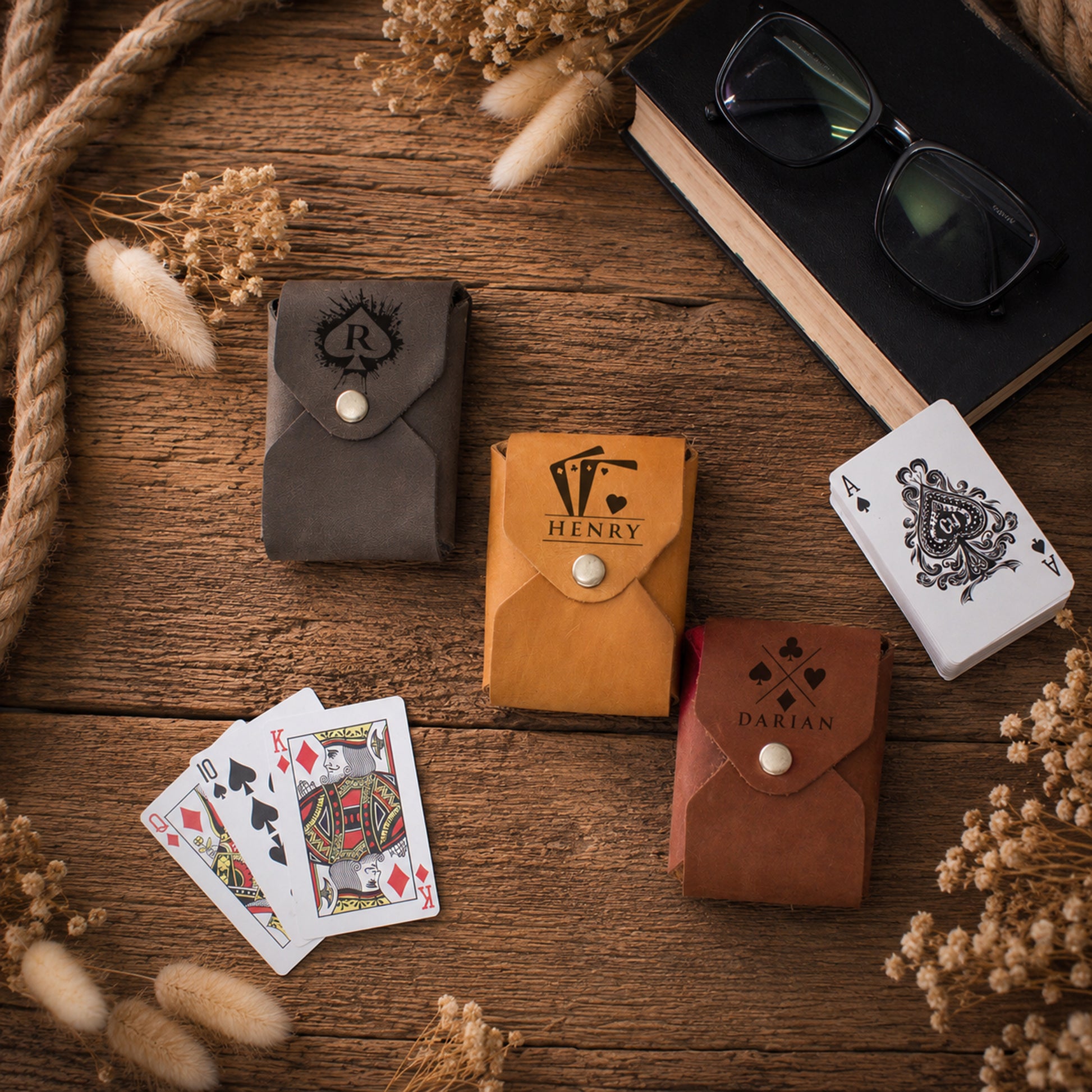 Four leather playing card sleeves in brown, tan, and dark gray are arranged on a rustic wooden table with playing cards, a book, and dried flowers. The tan sleeve features the brand name 'HENRY' printed on it.