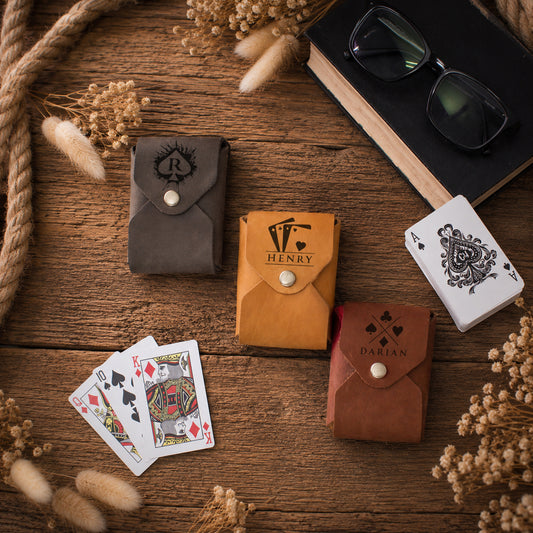 Four leather playing card sleeves in brown, tan, and dark gray are arranged on a rustic wooden table with playing cards, a book, and dried flowers. The tan sleeve features the brand name 'HENRY' printed on it.