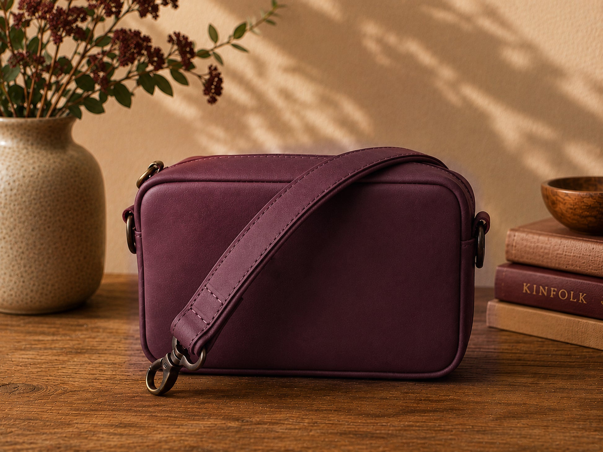 Burgundy crossbody bag on a wooden surface with a vase and books in the background