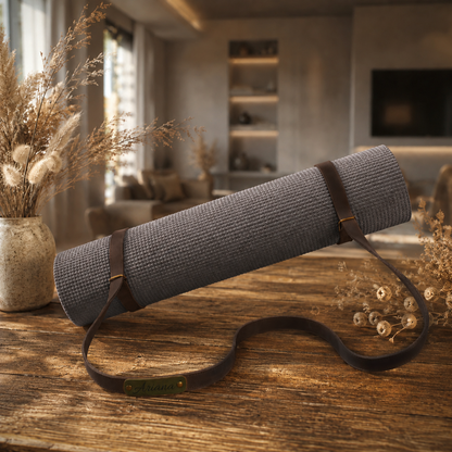 A gray textured yoga mat rolled up with a dark brown leather strap, resting on a rustic wooden table in a cozy, modern living room setting with dried flowers.
