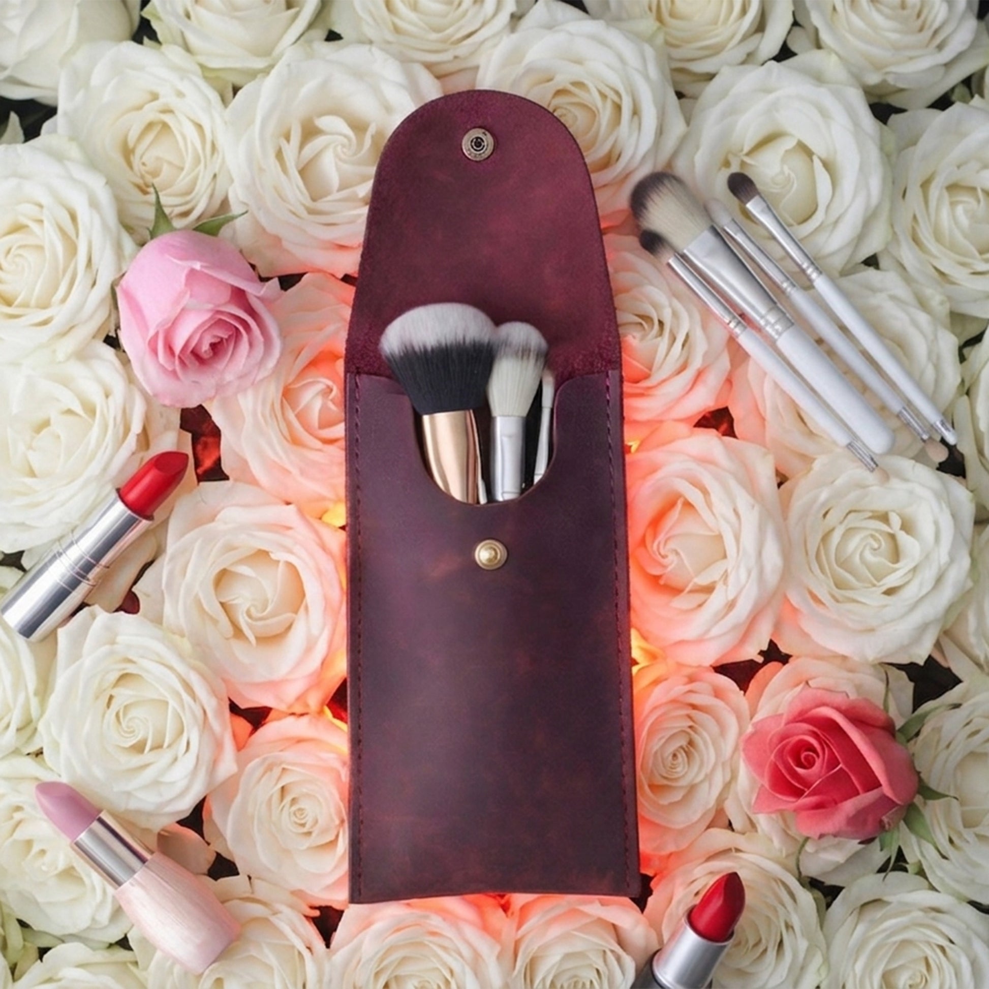 A dark burgundy leather makeup brush pouch with three makeup brushes inside, arranged on a bed of white and pink roses with lipstick props.