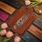 A set of personalized leather jewelry organizer pouches arranged on a rustic wooden surface with pink tulips and jewelry props. The central open brown pouch displays silver-toned necklaces and bracelets, while surrounding closed pouches in tan, black, magenta, gray, and blue show engraved names such as DAMARIS, Abigail, Frannie, K.