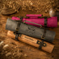 Three rolled canvas or fabric art/storage sleeves in gray, pink, and tan, each with dark leather straps and buckles, arranged on a wooden surface with dried flowers and rope props. The gray sleeve is most prominent and has a black monogram logo, while the tan sleeve at the bottom reads 'David Miller'.