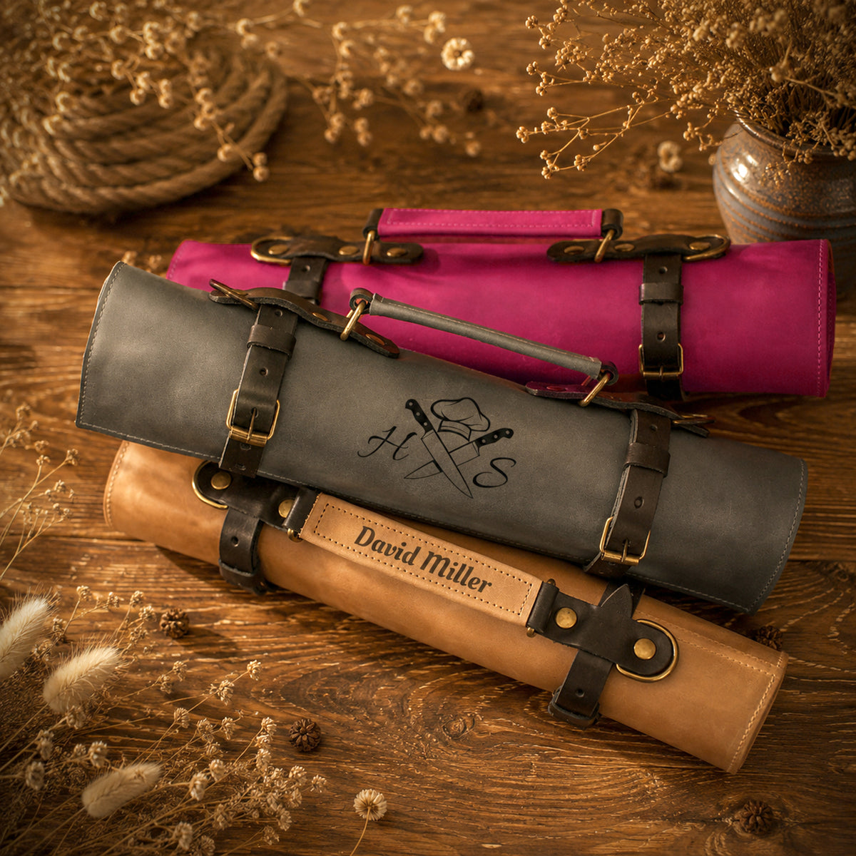 Three rolled canvas or fabric art/storage sleeves in gray, pink, and tan, each with dark leather straps and buckles, arranged on a wooden surface with dried flowers and rope props. The gray sleeve is most prominent and has a black monogram logo, while the tan sleeve at the bottom reads 'David Miller'.
