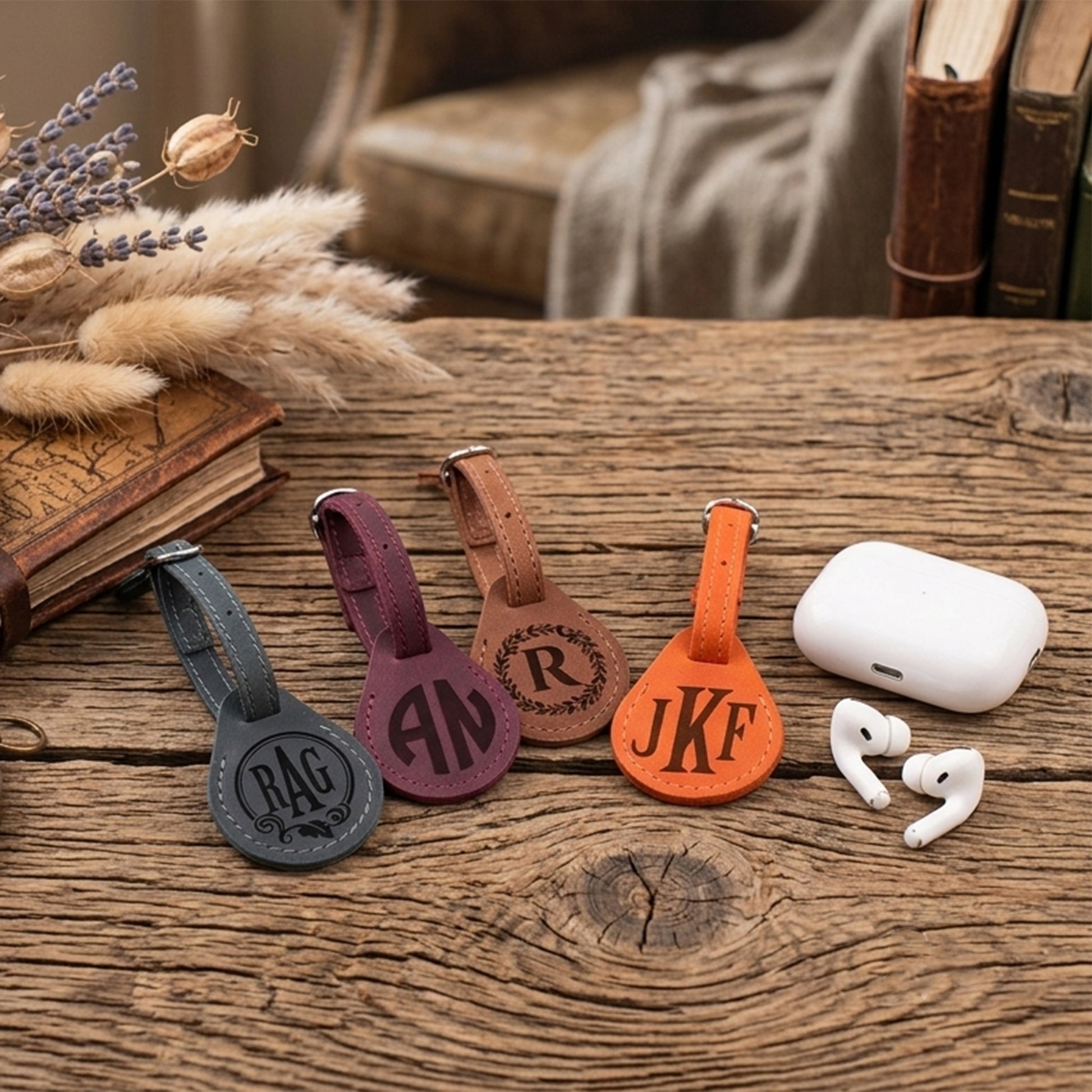 Four personalized leather keychains in gray, purple, brown, and orange, each engraved with a monogram or initials, arranged on a rustic wooden table next to a white wireless earbud charging case and earbuds.