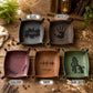 Five square ceramic dish holders with small side tabs, shown in different colors on a rustic wooden surface. Each dish has a black printed design and text: one says 'THE WORLD IS MYTY LQ' with a skyline, one says 'Jason' with a handprint, one says 'Regina' with floral script, one says 'I love you' with a soundwave, and one says 'WHEN YOU COME' with a dog silhouette.