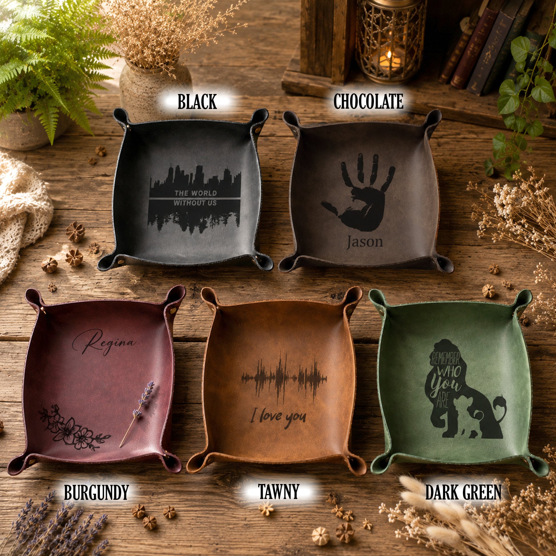 Five square ceramic dish holders with small side tabs, shown in different colors on a rustic wooden surface. Each dish has a black printed design and text: one says 'THE WORLD IS MYTY LQ' with a skyline, one says 'Jason' with a handprint, one says 'Regina' with floral script, one says 'I love you' with a soundwave, and one says 'WHEN YOU COME' with a dog silhouette.