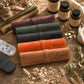 Five personalized leather pill organizer rolls tied with elastic bands, each labeled with a different name (TEDDY, Arthur, Jaxon, RILEY, Logan) in black script, arranged on a rustic wooden table alongside supplements, dried flowers, and books.