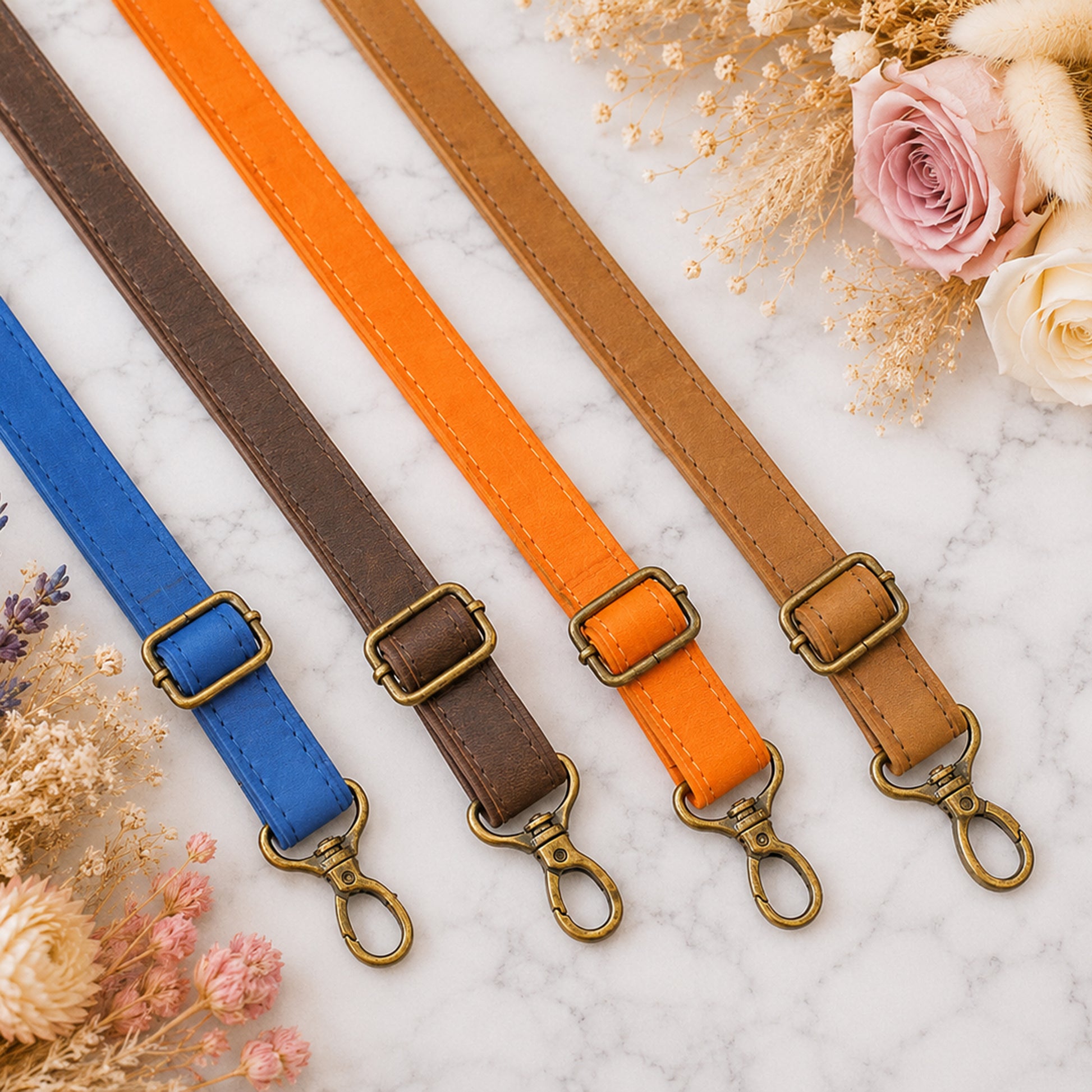 Four leather straps in blue, brown, orange, and beige with brass buckles on a light background with flowers.