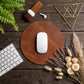 A round brown leather desk mat with a white wireless mouse on top, styled in a flat-lay on a dark wood tabletop with a keyboard, earbuds, dried flowers, and a gold geometric decor piece.