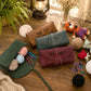 Three personalized crocheting or knitting tool rolls displayed on a wooden table with yarn balls, knitting needles, a lantern, and crochet props. The rolls are in brown, dark teal, and burgundy, with the names 'NELY', 'Coco', and 'Kirsten' embroidered on them.