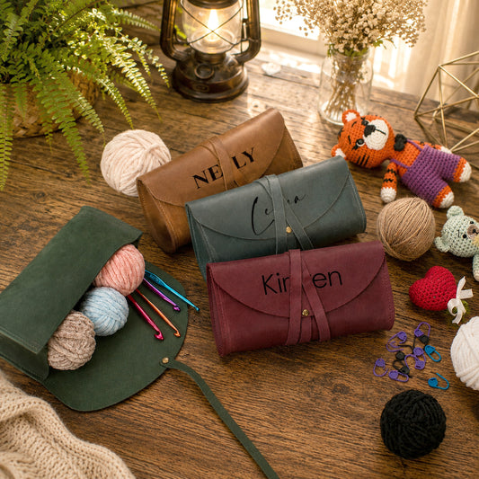 Three personalized crocheting or knitting tool rolls displayed on a wooden table with yarn balls, knitting needles, a lantern, and crochet props. The rolls are in brown, dark teal, and burgundy, with the names 'NELY', 'Coco', and 'Kirsten' embroidered on them.