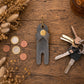 A gray leather key holder keychain with a gold snap button and brass keyring, displayed on a rustic wooden table next to keys and coins with dried flowers.