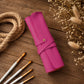 A rolled-up bright pink leather or faux-leather case with a snap strap and small gold-tone button, placed on a rustic wooden surface with paintbrushes, rope, and dried flowers around it.