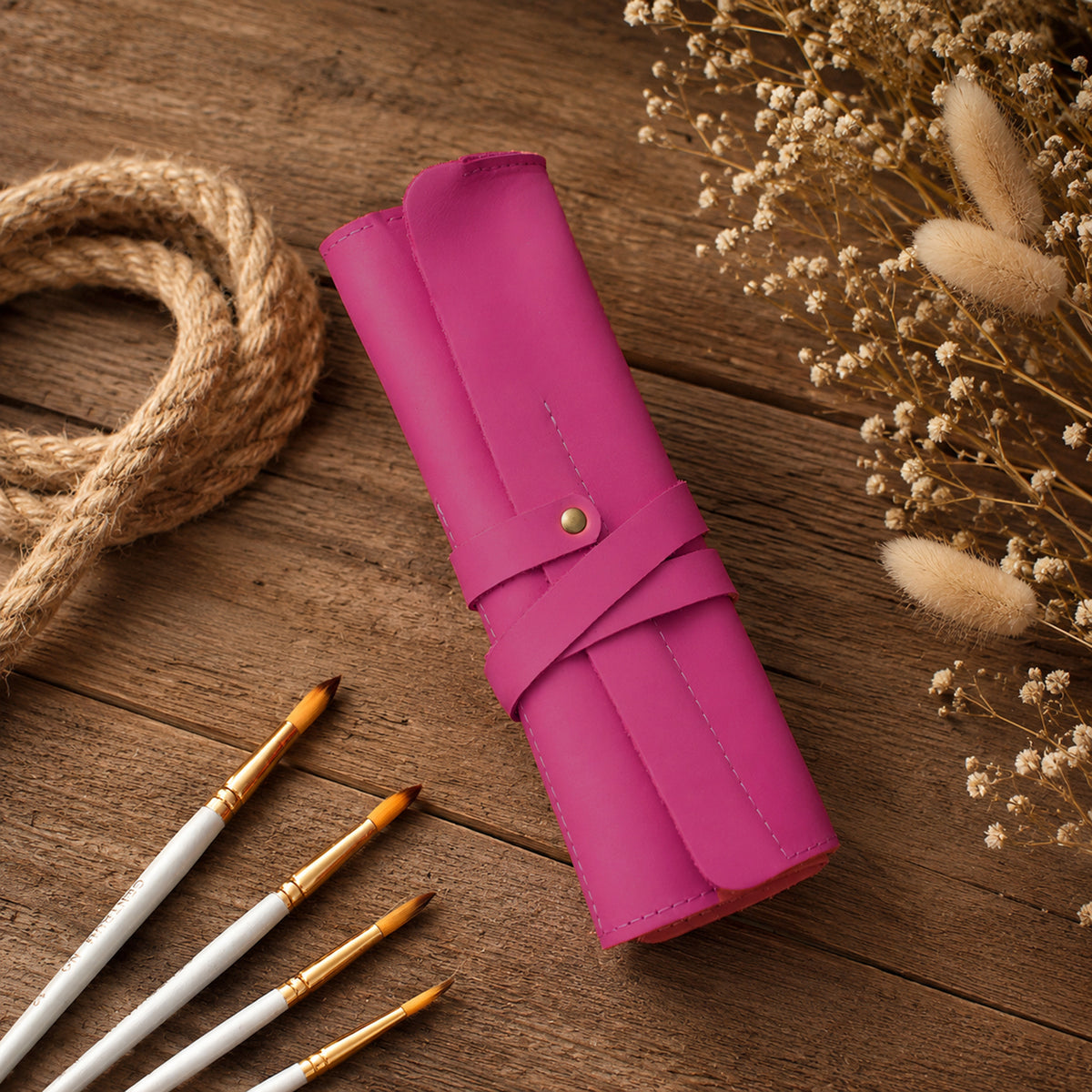 A rolled-up bright pink leather or faux-leather case with a snap strap and small gold-tone button, placed on a rustic wooden surface with paintbrushes, rope, and dried flowers around it.