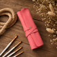 A pink leather roll-up case with a snap strap closure, styled on a rustic wooden surface with paintbrushes, rope, and dried flowers.