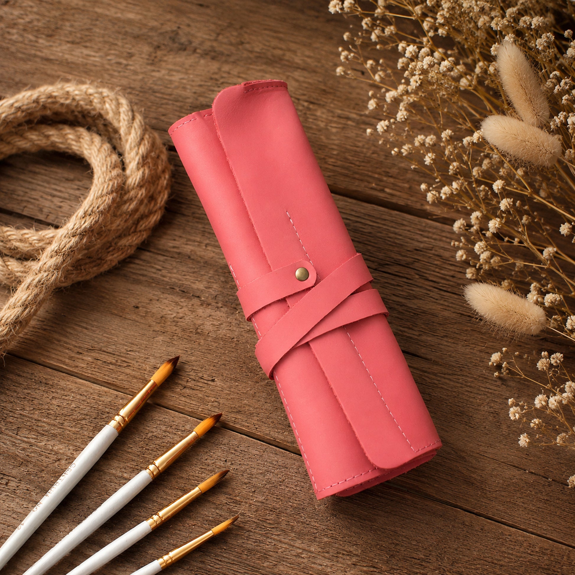 A pink leather roll-up case with a snap strap closure, styled on a rustic wooden surface with paintbrushes, rope, and dried flowers.