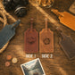 Two brown leather luggage tags shown on a wooden tabletop, labeled SIDE 1 and SIDE 2, with engraved mountain and letter designs. Surrounding props include a camera, rope, coins, a photo print, and other leather tags.