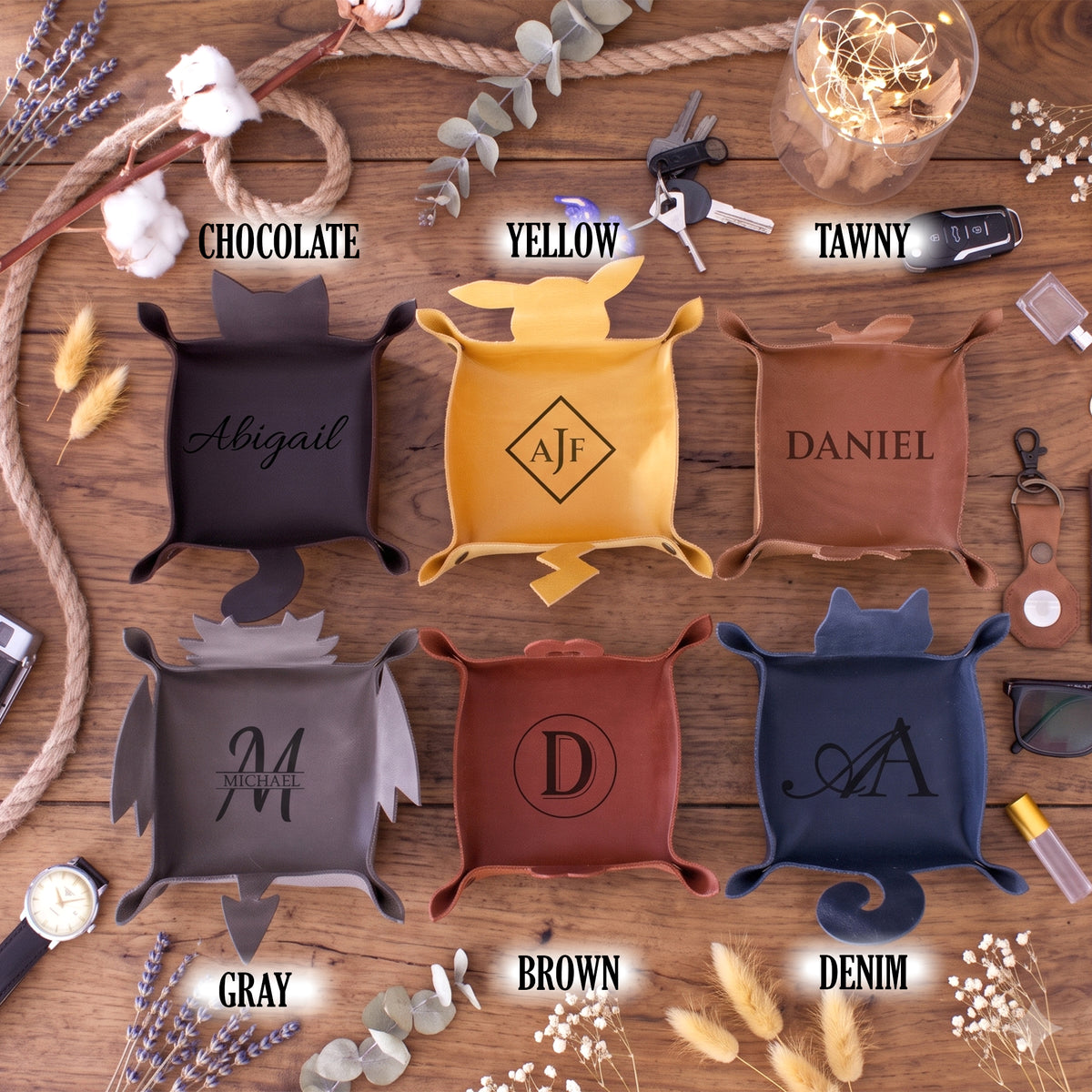 Six personalized cornhole bags in different colors (Chocolate, Yellow, Tawny, Gray, Brown, and Denim) arranged in a flat-lay on a rustic wooden table with decorative props like rope, cotton, and eucalyptus. Each bag has a different monogram or name printed on it.