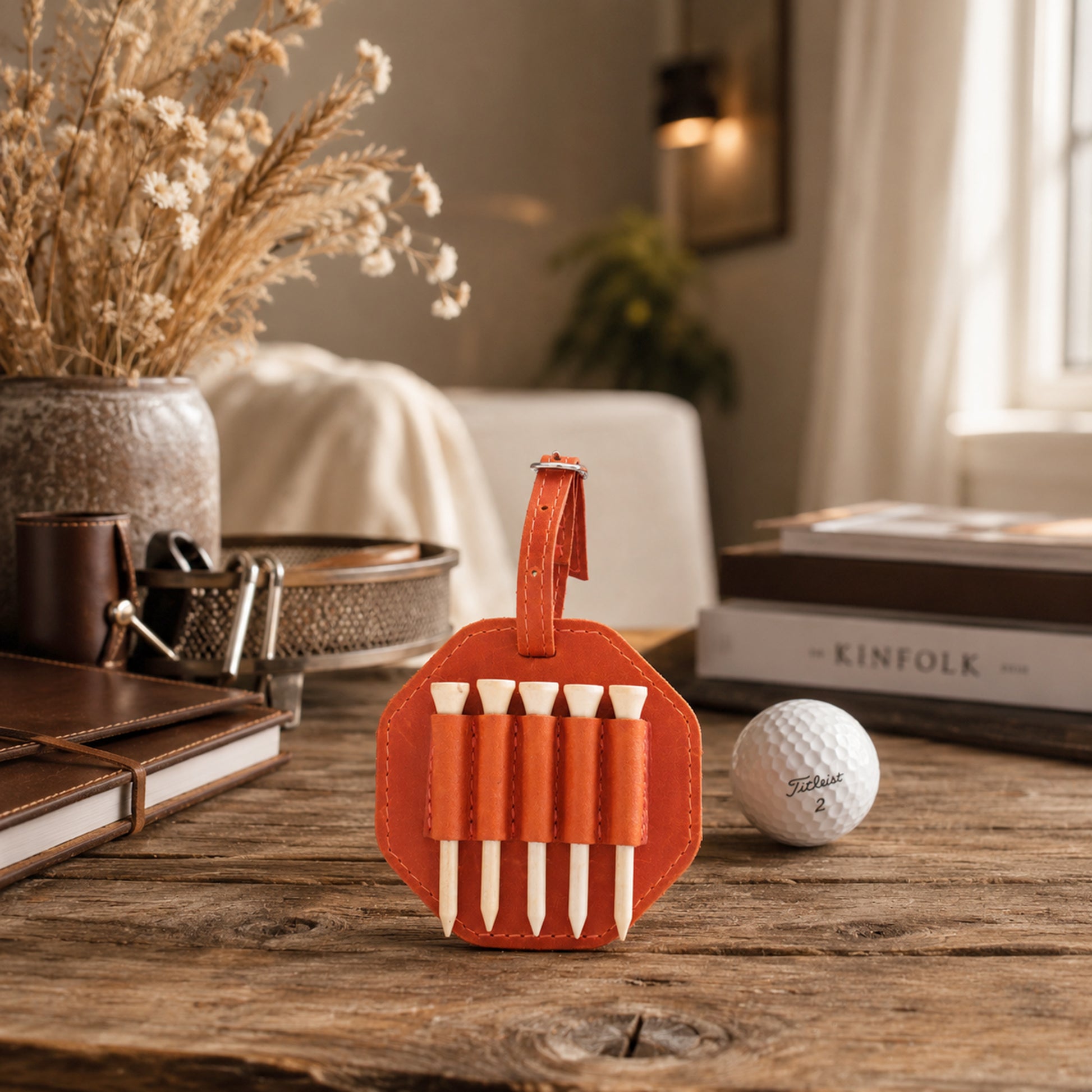 An orange leather golf tee holder with white tees, styled on a rustic wooden table next to a white golf ball, books, and dried flowers in a warm, cozy interior setting.