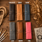 Six leather roll-up wallet or organizer cases arranged on a rustic wooden surface, each labeled with a color name: Chocolate, Tawny, Denim, Gray, Brown, and Pink. Decorative rope, dried flowers, paintbrushes, and a notebook are placed around them.