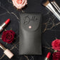 A black faux-leather makeup pouch with the name 'Della' in black script and a small pink pearl snap closure, styled on a black marble background with red roses, lipsticks, and makeup brushes.