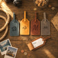 Four personalized leather luggage tags arranged on a rustic wooden table with travel-themed props, including a camera, rope, coins, a map, and instant photos. One tag is open, showing a blank name and address card insert.