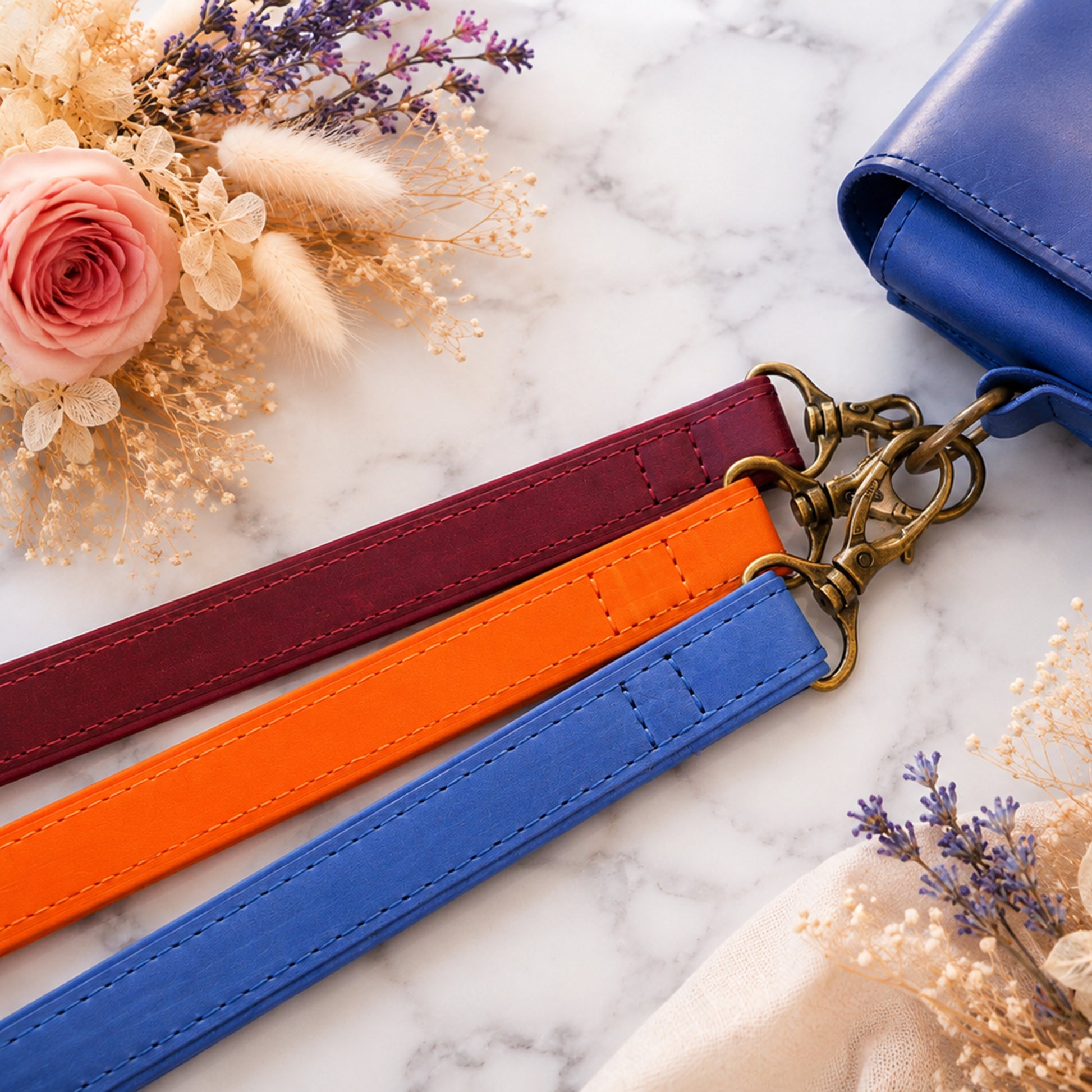 Three colorful leather straps (red, orange, blue) with a brass clasp on a marble surface with flowers.