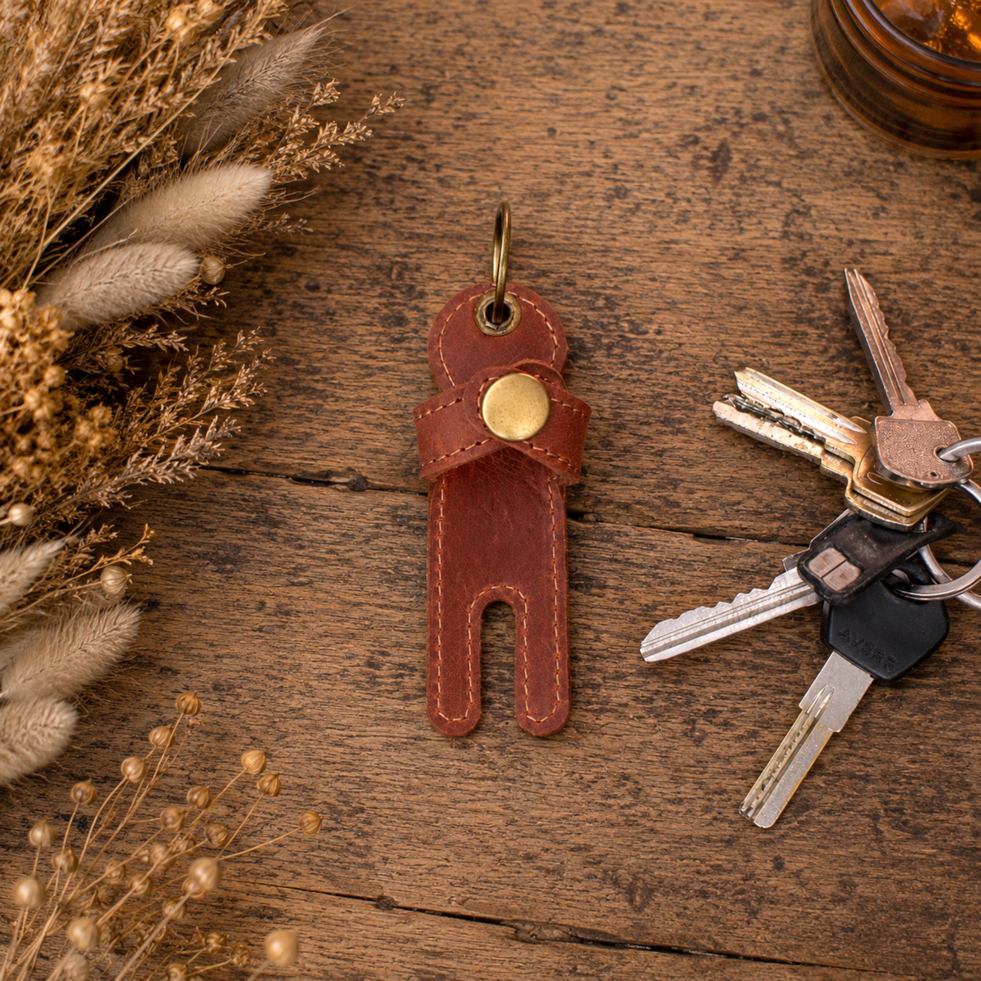 A brown leather key holder keychain with a gold snap button and metal key ring, displayed on a rustic wooden surface next to a set of keys and dried decorative grass.