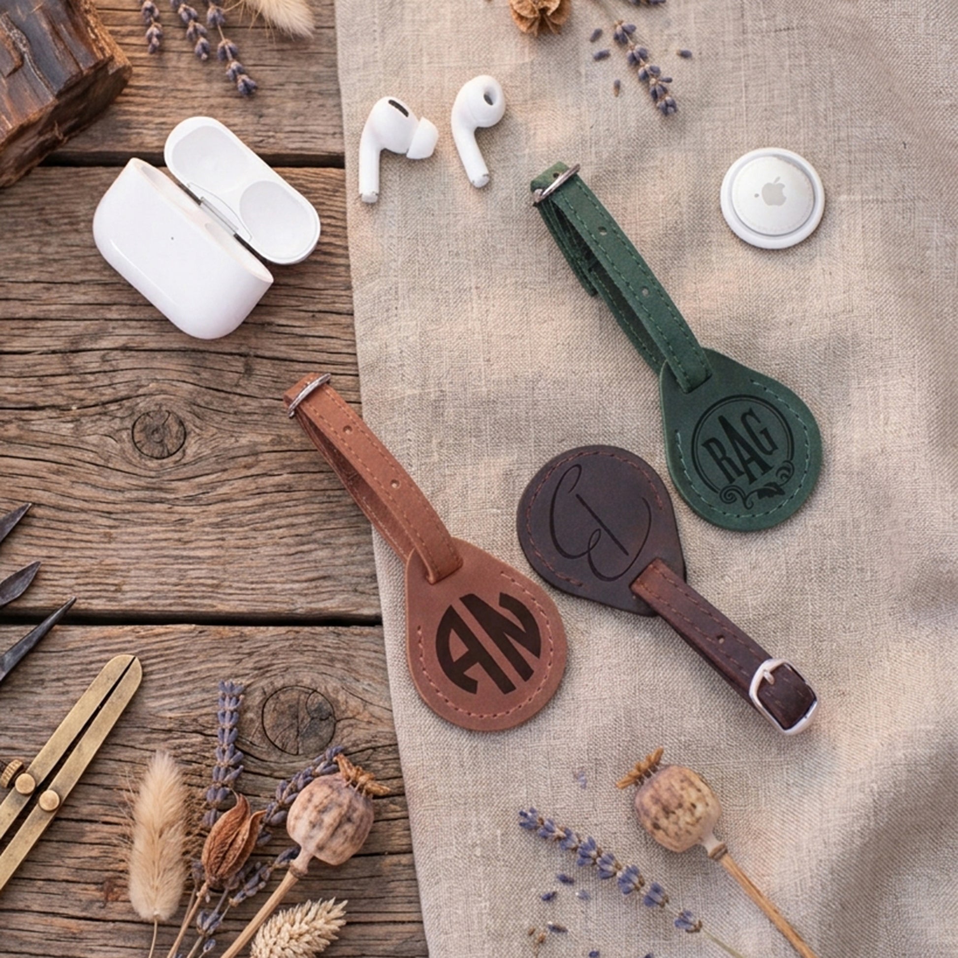 Three leather luggage tags in brown, dark brown, and green are arranged on a rustic wood and linen background with dried flowers, a white earbud case, and white wireless earbuds nearby.